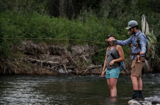 A fly fishing guide shows a female client how to cast while standing in a river
